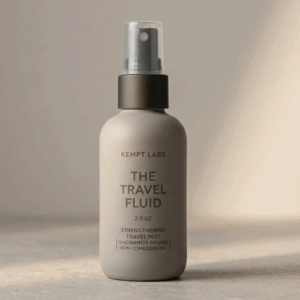 A travel size bottle of KEMPT Labs The Travel Fluid hair mist tucked into the front pocket of a dark grey gym bag.
